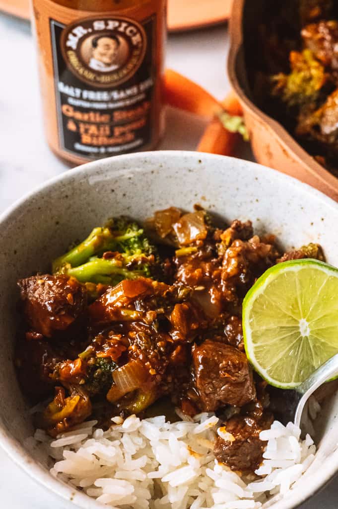 Beef and Broccoli in a bowl with a lime wedge and rice surrounded by Mr. spice bottle and pink pan