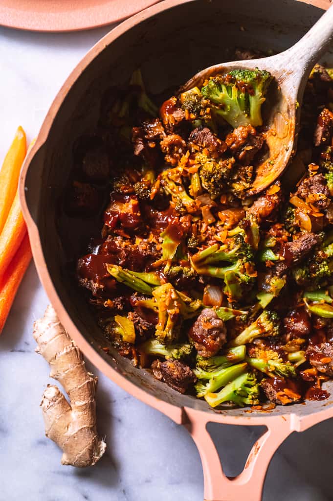 Beef and broccoli in a pan with a wooden spoon next to carrots and ginger