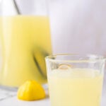 Glass of lemonade in front of a pitcher and slice of lemon
