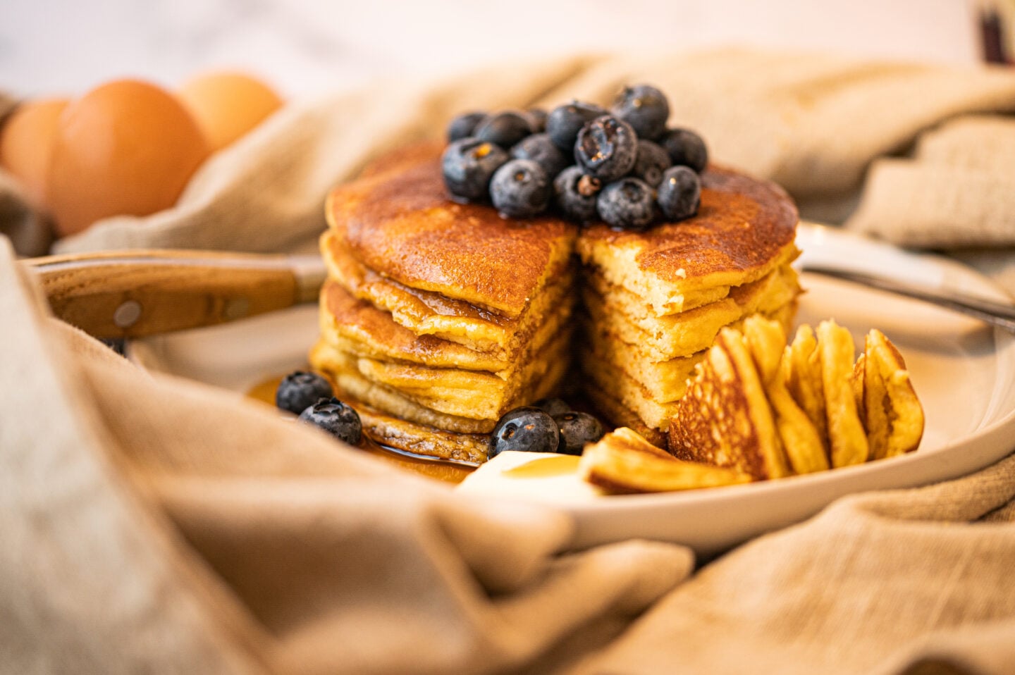 Golden almond‑flour pancakes stacked tall, covered in syrup and fresh blueberries, styled in a cozy kitchen setting