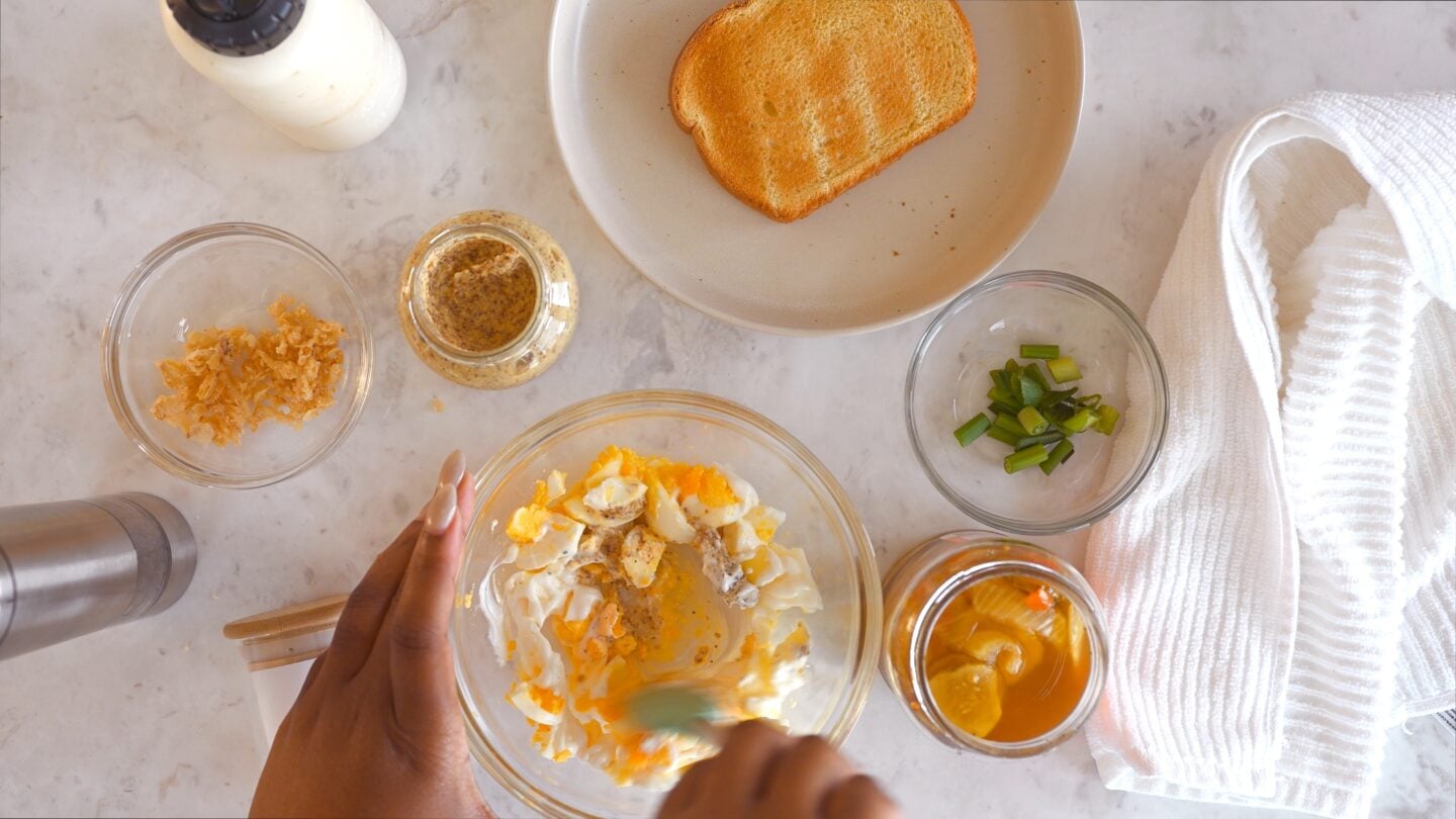 Flat lay of egg salad ingredients including bread, mayo, mustard, french fried onions, green onions, pickle juice, eggs, salt and pepper.