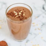 Glass filled with a creamy chocolate-banana smoothie topped with rolled oats and cocoa powder, placed on a marble countertop with scattered oats and cocoa powder in the background