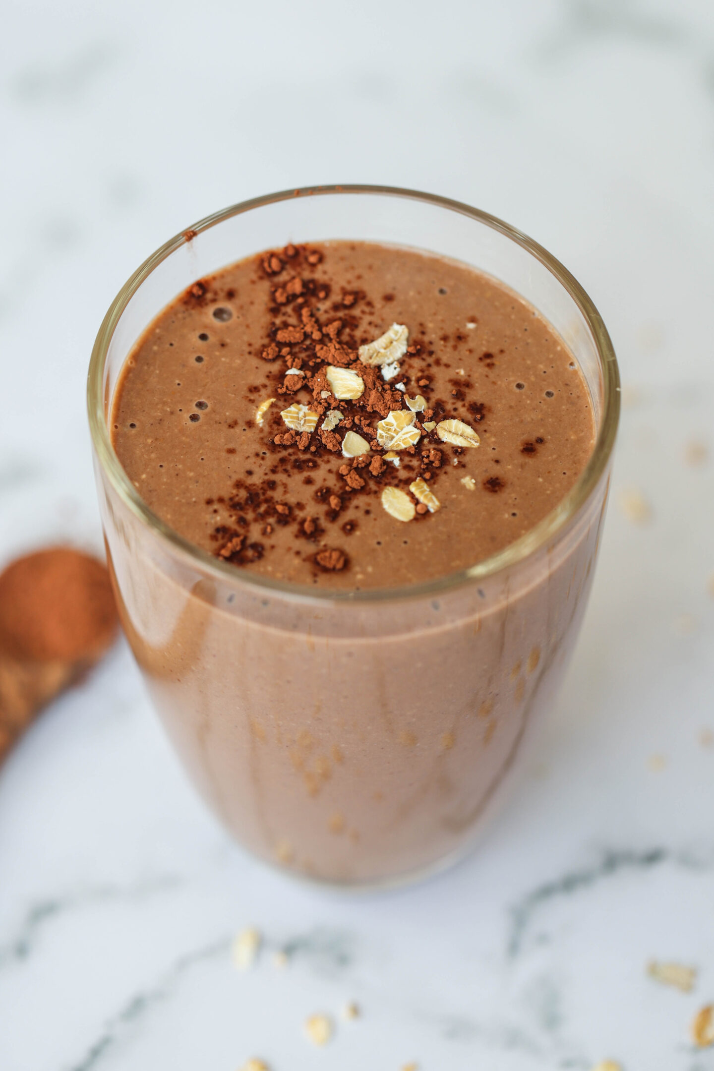 Glass of creamy chocolate-banana coffee smoothie topped with oats and cocoa powder, placed on a light marble surface with scattered oats and cocoa in the background