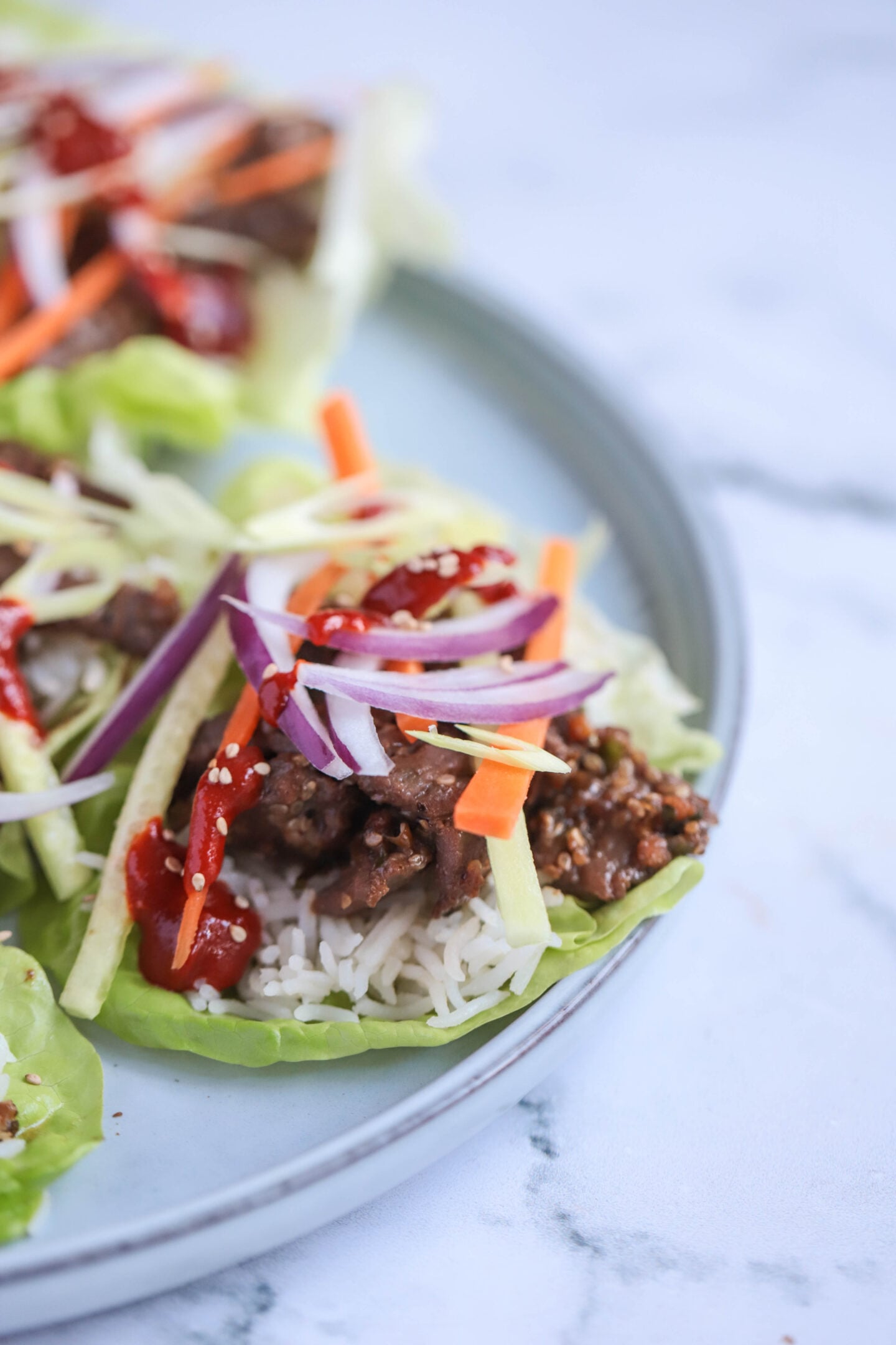 "Plate of Korean beef bulgogi lettuce wraps featuring tender marinated beef, fluffy white rice, and crisp matchstick vegetables, topped with sliced red onions, sesame seeds, and a drizzle of gochujang sauce, served on fresh green lettuce leaves against a marble background."