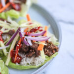 Plate of Korean beef bulgogi lettuce wraps featuring tender marinated beef, fluffy white rice, and crisp matchstick vegetables, topped with sliced red onions, sesame seeds, and a drizzle of gochujang sauce, served on fresh green lettuce leaves against a marble background.