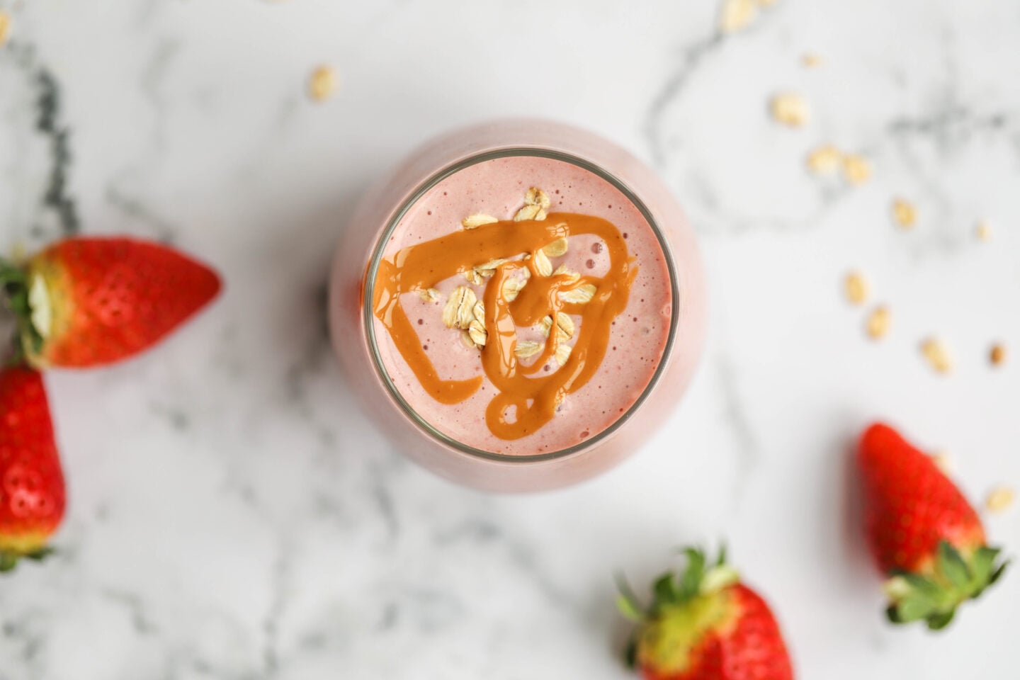 A close-up top-down view of a strawberry smoothie in a glass. The smoothie is decorated with peanut butter drizzle and oats. Fresh strawberries and oats are spread across a white marble background.
