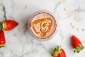 A close-up top-down view of a strawberry smoothie in a glass. The smoothie is decorated with peanut butter drizzle and oats. Fresh strawberries and oats are spread across a white marble background.
