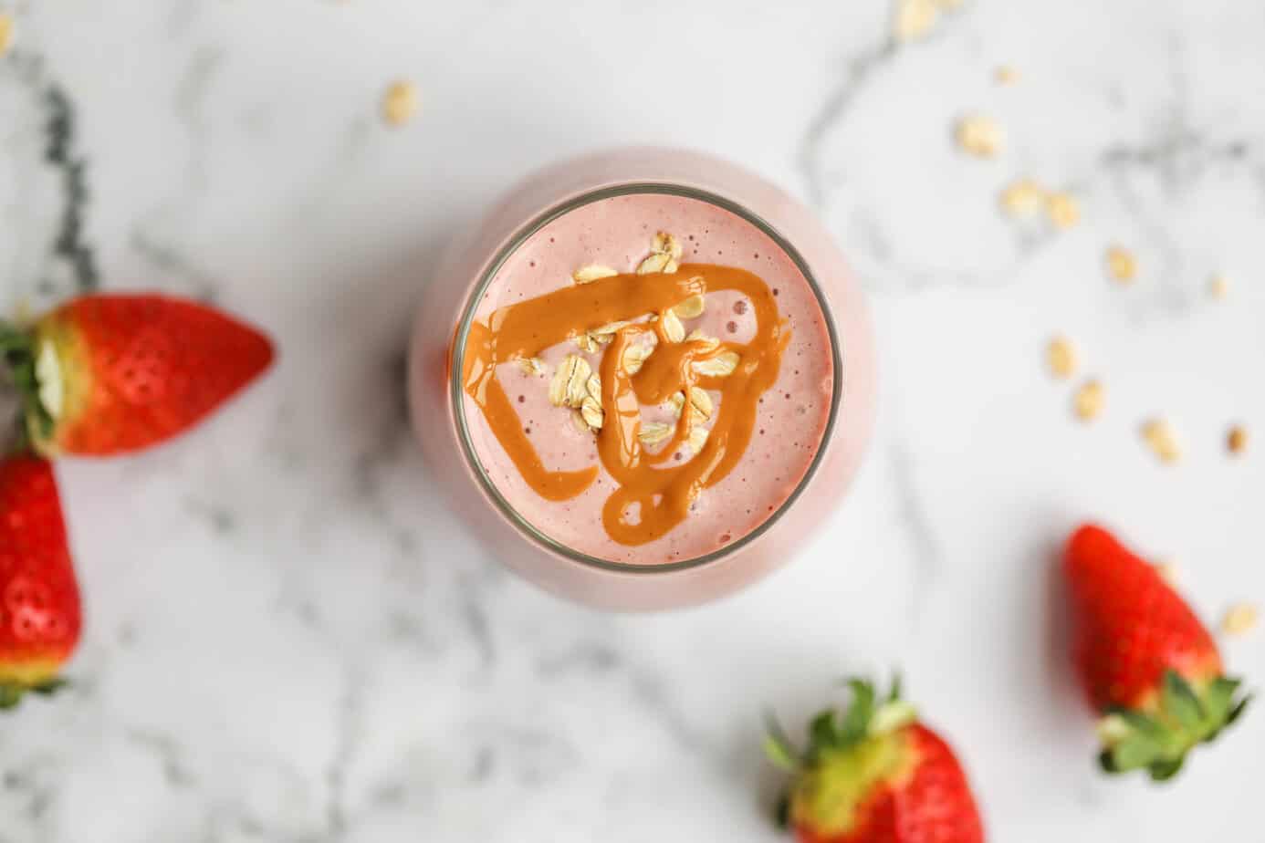 A close-up top-down view of a strawberry smoothie in a glass. The smoothie is decorated with peanut butter drizzle and oats. Fresh strawberries and oats are spread across a white marble background.