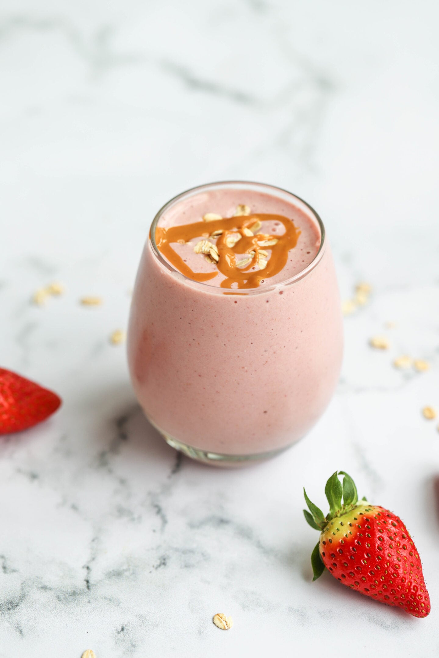 A side view of a strawberry smoothie in a clear glass on a white marble surface. The smoothie is light pink, topped with a drizzle of peanut butter and oats, with fresh strawberries placed on the side for decoration.