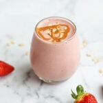 A side view of a strawberry smoothie in a clear glass on a white marble surface. The smoothie is light pink, topped with a drizzle of peanut butter and oats, with fresh strawberries placed on the side for decoration.