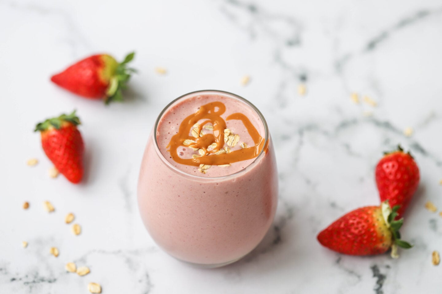  wide-angle shot of a strawberry smoothie in a glass, surrounded by fresh strawberries and oats on a white marble surface. The smoothie is light pink with a peanut butter drizzle and oat topping.