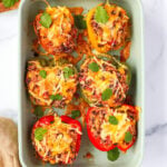 A baking dish filled with colorful quinoa stuffed bell peppers, topped with melted cheese and garnished with fresh cilantro. The peppers are red, yellow, and green, with a hearty filling of quinoa, black beans, corn, and spices. A wooden cutting board with extra cilantro is visible in the background.