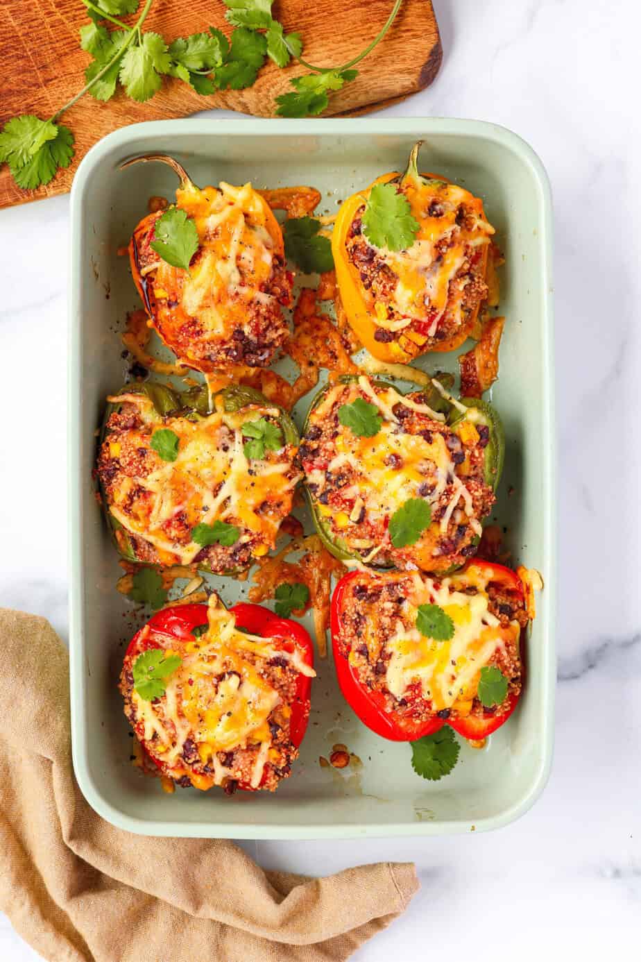 A baking dish filled with colorful quinoa stuffed bell peppers, topped with melted cheese and garnished with fresh cilantro. The peppers are red, yellow, and green, with a hearty filling of quinoa, black beans, corn, and spices. A wooden cutting board with extra cilantro is visible in the background.