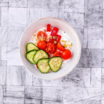 Overhead shot of a fresh cottage cheese bowl with cucumber slices, halved cherry tomatoes, red bell pepper, and a sprinkle of pistachios, arranged on a textured gray surface.