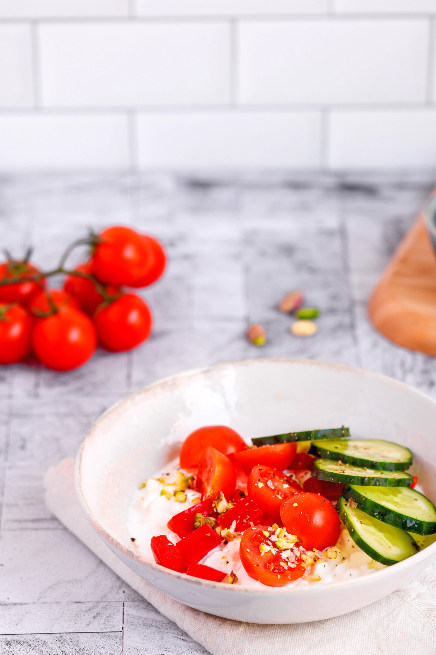 Top-down view of a savory cottage cheese bowl topped with sliced cucumber, cherry tomatoes, red bell pepper, and chopped pistachios, with a small bowl of pistachios on a wooden board nearby.