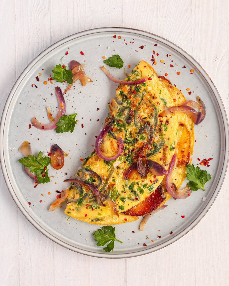 Top-down view of a folded potato cheese omelet garnished with red onion, chopped parsley, and chili flakes on a gray plate — a high-protein, diabetes-friendly breakfast option.